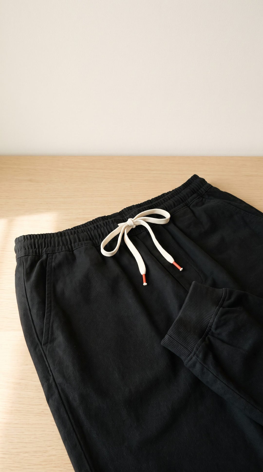 Close-up of the black harem trousers' waist on a pale oak table: cream drawstring with orange-red tips