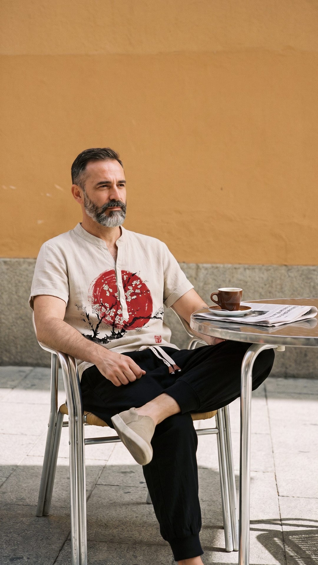 Man around 48 with a salt-and-pepper beard at a café table, wearing a red sun-print shirt and black harem trousers