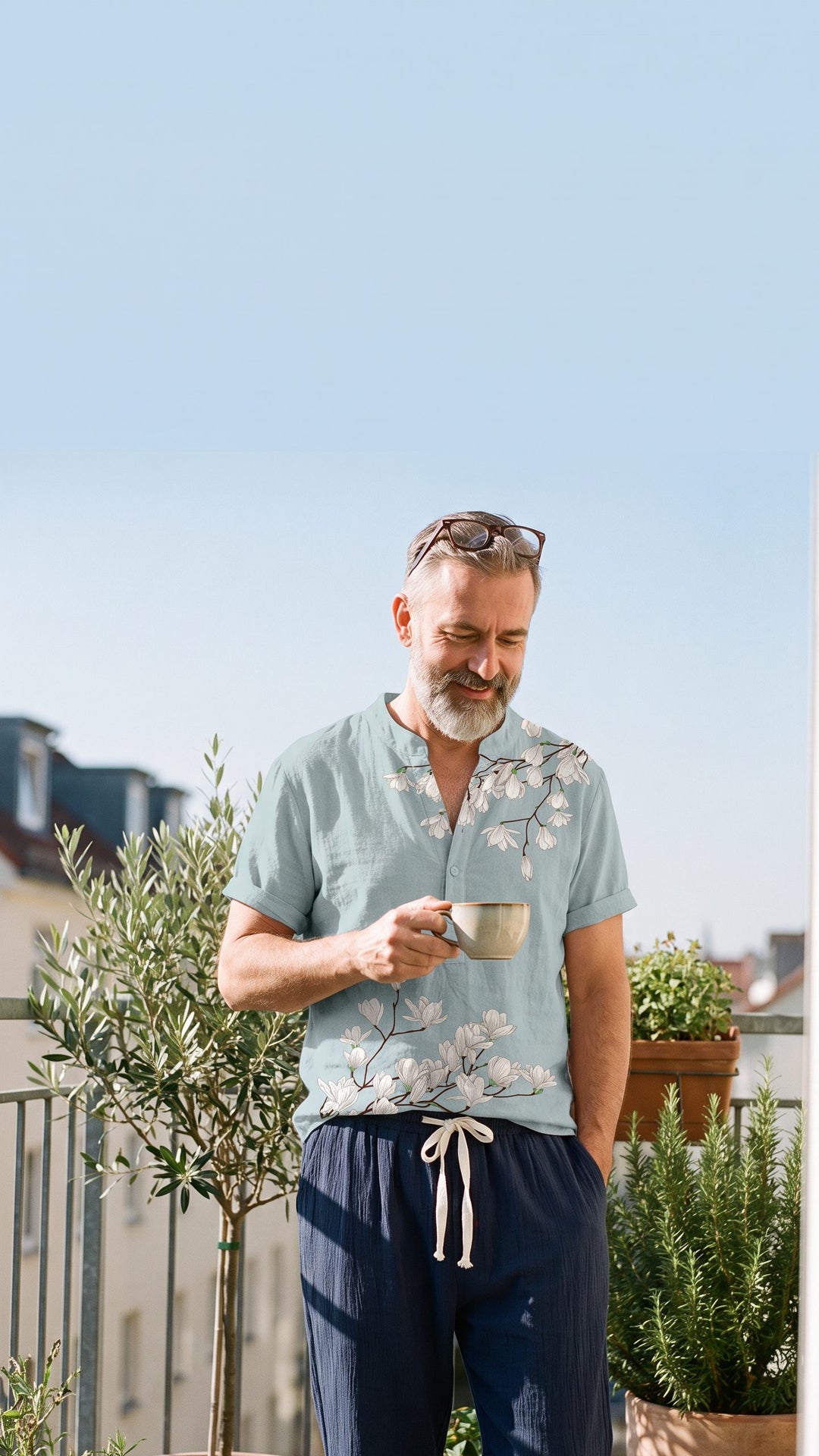 Man around 52 with a grey beard on a sunny balcony in a blue cherry-blossom print shirt and navy trousers