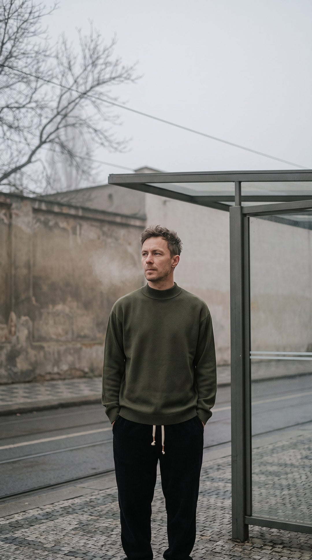 Man around 40 waiting at a misty tram stop in black corduroy trousers and an army-green sweatshirt